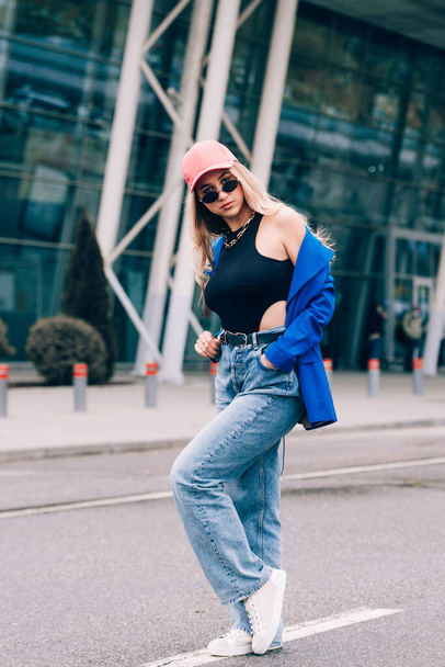 Young sexy blonde hipster woman posing on the street. Wearing blue stylish jacket, jeans and baseball hat and sunglasses. - Photo, Image