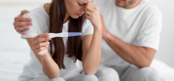 Обрізаний розчарований сумний молодий кавказький чоловік підтримує свою дружину, проводить тест на вагітність, сидить на ліжку в спальні
 - Фото, зображення