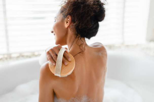 Back view of young woman making lymphatic massage with brush, scrubbing her skin in foamy bath at home - Photo, Image