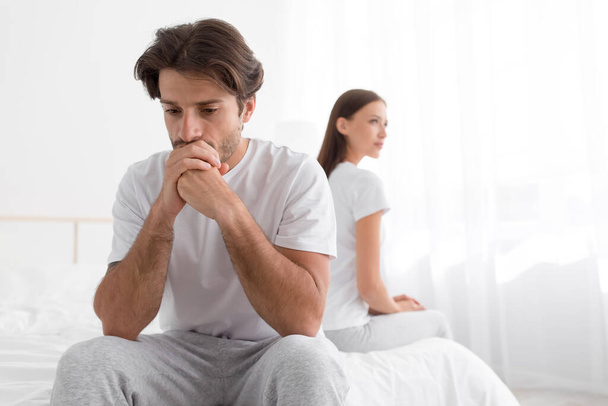 Sad upset offended millennial caucasian man sitting on bed, ignoring woman in bedroom interior - Zdjęcie, obraz
