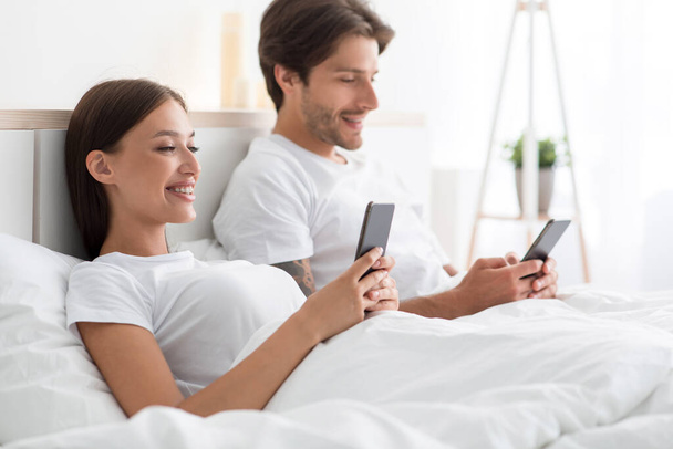 Fröhliche junge kaukasische Männer und Frauen sehen sich Videos an oder chatten in sozialen Netzwerken in Smartphones im Bett - Foto, Bild