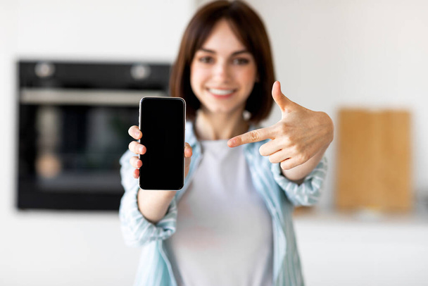 Senhora feliz mostrando tela de smartphone em branco e apontando sobre ele enquanto está em pé no interior da cozinha, foco seletivo - Foto, Imagem
