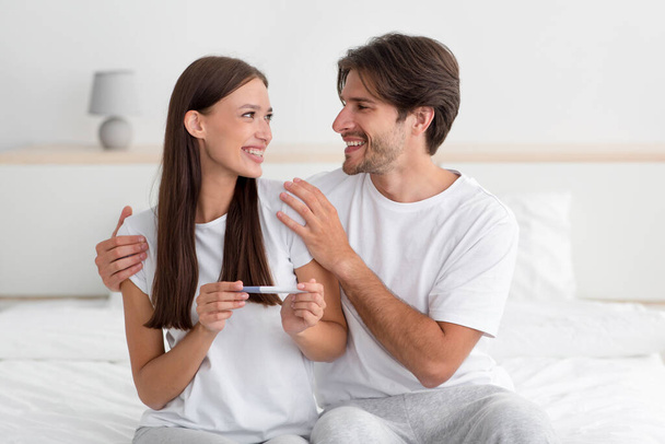 Lächelndes europäisches Ehepaar freut sich über Ergebnis des Schwangerschaftstests, sitzt im Bett - Foto, Bild