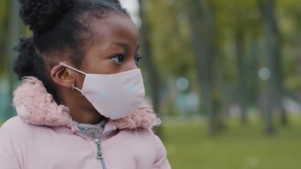 Tıbbî maskeli küçük kız, yüzünü çevirmiş, kameraya bakıyor. Park pandemik koronavirüs karantinasında poz veren maskeli çocuk öğrenci öğrenci portresi - Video, Çekim