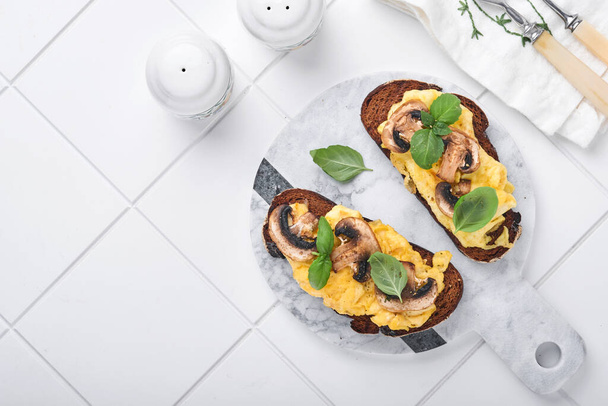 Scrambled eggs with fried mushrooms and basil on bread on white table background. Homemade breakfast or brunch meal. Scrambled eggs and mushrooms sandwiches. Top view with copy space - Photo, image