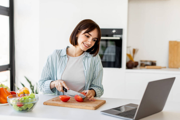 Νεαρή γυναίκα μαγειρεύει φρέσκια βιολογική σαλάτα, κόβει ντομάτες και κοιτάζει το laptop στο πάγκο, δοκιμάζοντας νέα συνταγή - Φωτογραφία, εικόνα