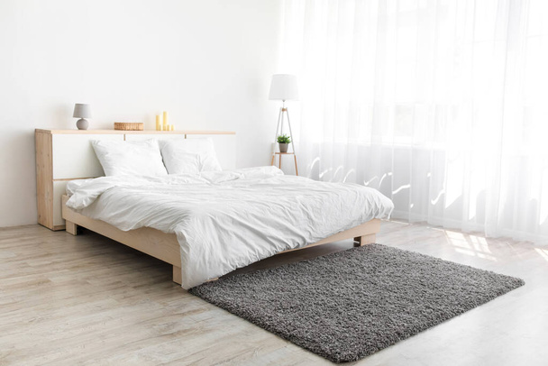 Большая кровать с удобными постельными принадлежностями, мягкий серый ковер, лампа на полу, мебель с аксессуарами - Фото, изображение