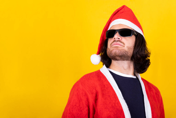 Молодой человек в костюме Санта-Клауса носит длинные волосы и солнцезащитные очки. Желтый фон и место для копирования сообщения. - Фото, изображение