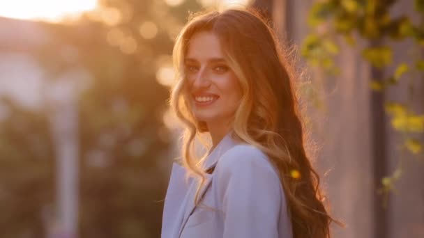 Portrait beautiful attractive happy Caucasian girl with long hair stands sun rays background looking at camera smiling sincerely toothy with white teeth. Millennial girl posing in sunlight with smile - Footage, Video