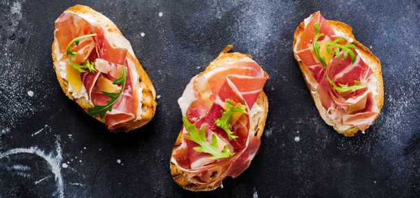 Open sandwiches with jamon, arugula and hard cheese on a concrete old dark background. Rustik style.Top view. - Foto, afbeelding
