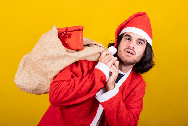 Joulupukiksi pukeutunut nuori mies kantaa säkillistä lahjoja. Hänellä on pitkät hiukset, parta ja yllätyksen ilme. Kuva eristetty keltaisella pohjalla. - Valokuva, kuva