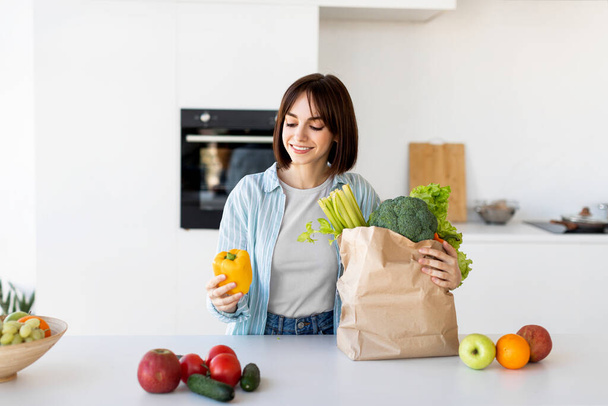 Jeune dame déballer sac d'épicerie après les achats, mettre des légumes sur la table dans la cuisine intérieure à la maison, copie épicée - Photo, image