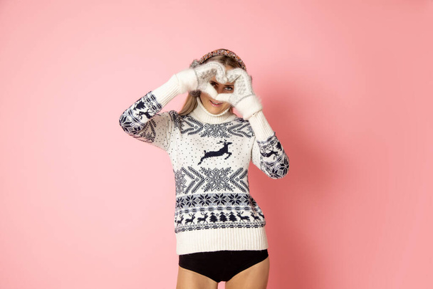 Portrait of cute pretty woman in shorts 30-35 years in sweater showing heart with hands in gloves. Studio shot on pink background. Concept of personal accessories . Fashion model. winter clothing. - Photo, Image