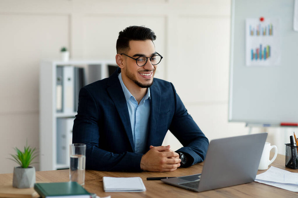 Αυτοπεποίθηση νεαρός Άραβας επιχειρηματίας με επίσημο κοστούμι χρησιμοποιώντας φορητό υπολογιστή στο γραφείο του, έχοντας εικονική συνάντηση στο γραφείο - Φωτογραφία, εικόνα