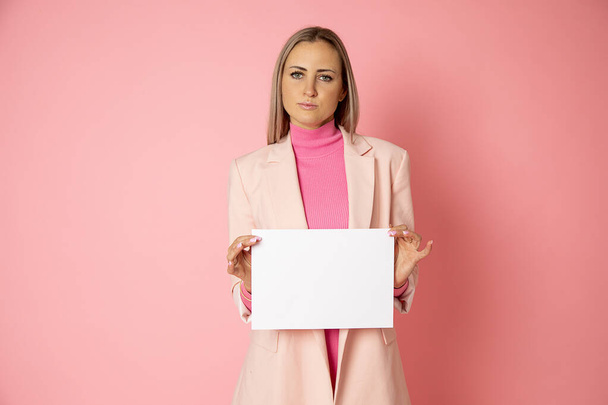 Portret atrakcyjnej młodej blondynki kobiety 30-35 lat trzymającej pusty papier A4 na różowym tle. Bizneswoman ma na sobie różową kurtkę. Najlepszy menedżer, blog kobiet - Zdjęcie, obraz