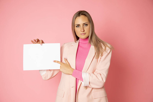 Portret van blond haar aantrekkelijke vrouw 35-40 jaar houden blanco van papier A4 voor teken op roze achtergrond. Zakenvrouw draagt een roze jasje. Punten met vinger op kopieerruimte. - Foto, afbeelding