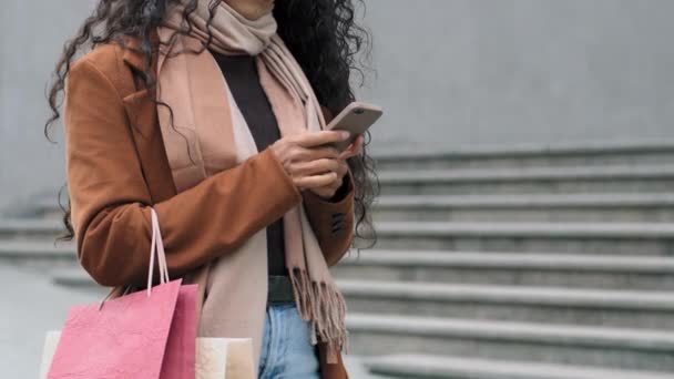 クローズアップ撮影認識できない若いスタイリッシュな女の子の買い物客の消費者の女性は、携帯電話オンラインショッピングアプリでチャット購入と明るいバッグを保持街の通りに立って - 映像、動画