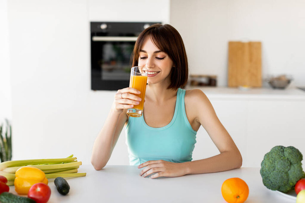 Dieta odwykowa. Slim lady picia soku pomarańczowego podczas gotowania zdrowej kolacji warzywnej, siedzi w jasnonowoczesnej kuchni - Zdjęcie, obraz