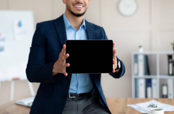 Nierozpoznawalny arabski biznesmen pokazujący tablet z czystym ekranem, oferujący makietę na stronę internetową lub aplikację w biurze - Zdjęcie, obraz