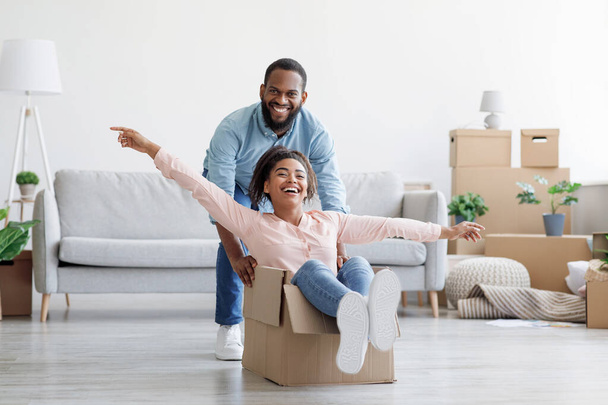 Satisfecho riendo joven afroamericano marido empujando caja con mujer emocionada en nuevo apartamento en sala de estar - Foto, Imagen