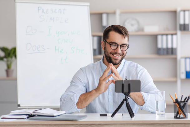 Amigável adulto europeu professor masculino em óculos com quadro-negro, gestos, olha para smartphone webcam - Foto, Imagem