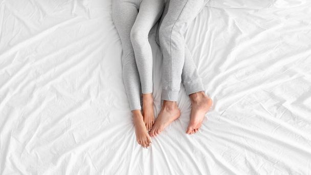 Legs of caucasian european husband and wife lie together on white bed in bedroom, top view - Photo, image
