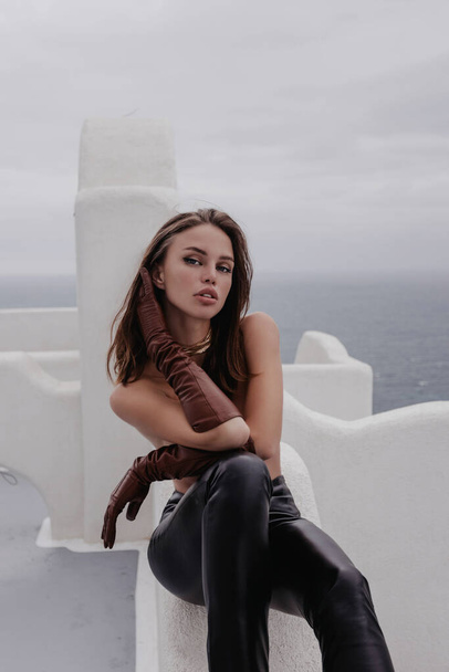 fashion outdoor photo of beautiful woman with dark hair in elegant leather clothes posing at Santorini  - Fotografie, Obrázek