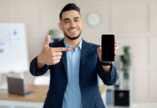 Молодой арабский бизнесмен указывает на смартфон с чистым экраном в современном офисе, макет для мобильного приложения или сайта - Фото, изображение