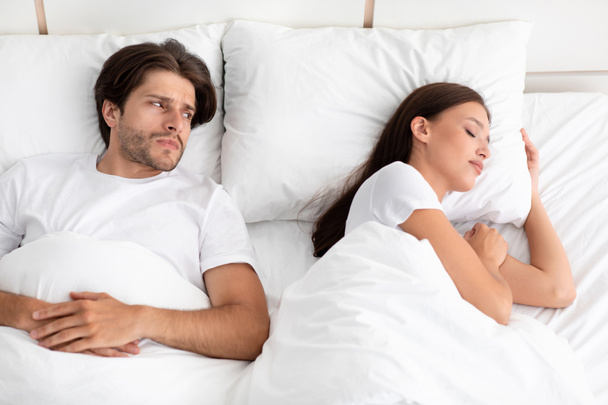 Disgruntled sad caucasian young guy does not sleep, lies in bed, looks at lady, woman snores in white bedroom - Photo, image