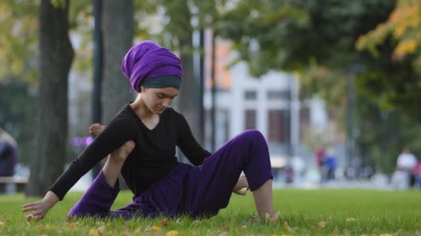 Girl in hijab muslim woman sports lady female yogi sitting in city park on grass lawn holds foot under arm doing stretching grip with hands yoga practice training stretching outdoors sport exercises - Footage, Video