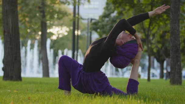 Muslim islamic girl in hijab trainer guru yoga master woman doing stretching exercises in park on green grass doing sports sits in beautiful acrobatic pose asana with back bend stretch on lawn outdoor - Footage, Video