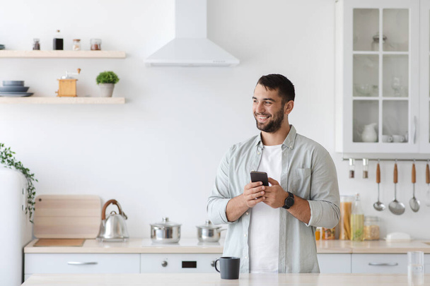Happy adult caucasian guy with smartphone and cup of coffee look at empty space at kitchen interior - Photo, image