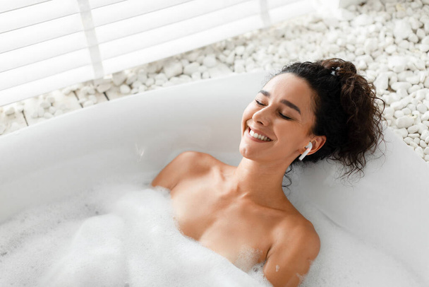 Calma sorridente giovane donna rilassante in bagno di bolle con gli occhi chiusi, ascoltare musica o meditazione audio in auricolare - Foto, immagini