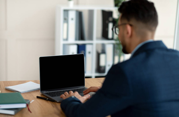 Nerozpoznatelný arabský podnikatel pracující na notebooku s prázdnou obrazovkou v kanceláři, maketa pro design webových stránek - Fotografie, Obrázek
