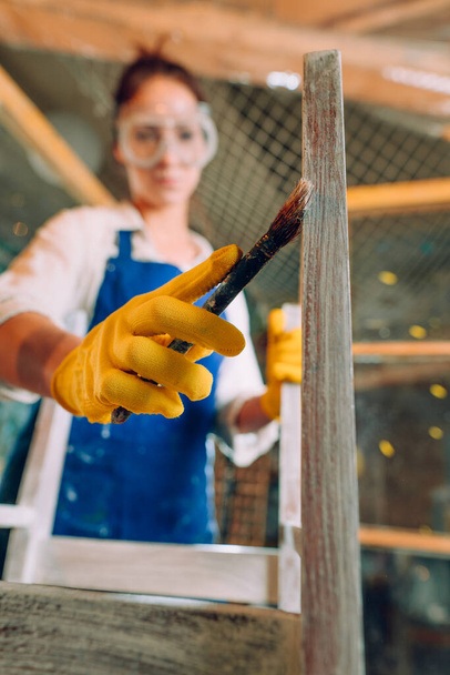 Woman renovating a chair using a paintbrush. Young woman wearing protective glasses and apron working in the workshop. Selective focus on the paintbrush. Upcycling concept. - Φωτογραφία, εικόνα