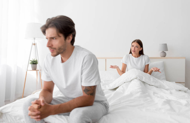 Расстроенный молодой европейский мужчина, сидящий на кровати, игнорируя леди. Жена кричит и ссорится в спальне - Фото, изображение