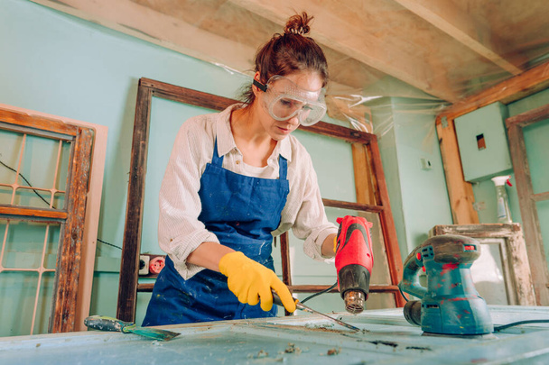 Egy nő, aki leveszi a festéket az ajtóról egy ipari hajszárítóval. A műhelyben dolgozó fiatal nő védőszemüveget, kesztyűt és kötényt visel. Újrahasznosítási koncepció. - Fotó, kép