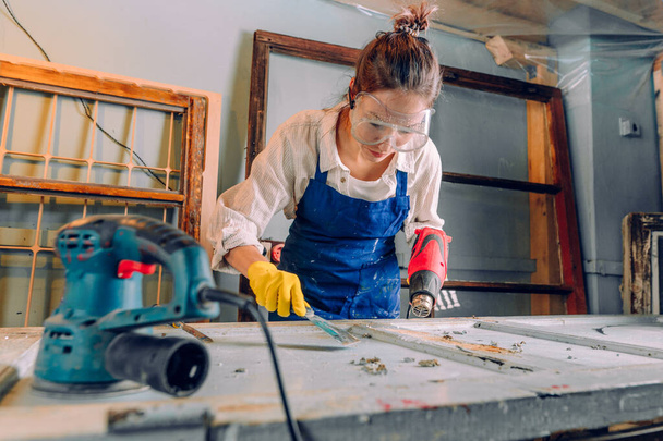 Женщина снимает краску с двери, используя промышленный фен. Молодая женщина в защитных очках, перчатках и фартуках работает в мастерской. Концепция подъема. - Фото, изображение