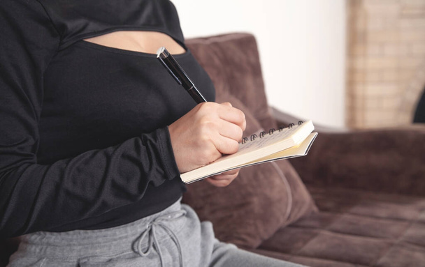Młoda kobieta siedzi na kanapie pisze w notatniku. - Zdjęcie, obraz