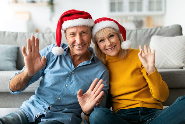 Senior pár Waving Hello Noaring Santa Klobouky Slaví Vánoce Indoor - Fotografie, Obrázek