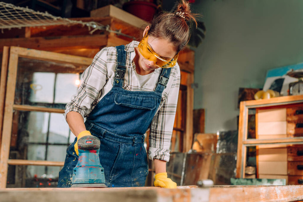 Retrato de una mujer con gafas protectoras y guantes que trabaja en el taller con una lijadora de discos. Concepto de Upcycling. - Foto, Imagen