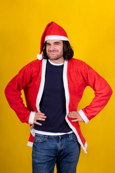 Счастливый молодой человек одет как Санта Клаус. Мальчик держит руки на бедрах и смотрит в камеру. Желтый фон и пространство для копирования. - Фото, изображение