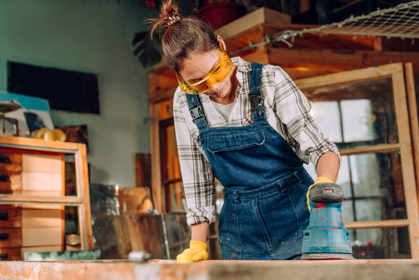 Retrato de una mujer con gafas protectoras y guantes que trabaja en el taller con una lijadora de discos. Concepto de Upcycling. - Foto, Imagen