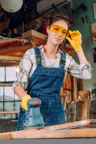 A portrait of a woman wearing protective glasses and gloves working in the workshop using a disc sander. Upcycling concept. - Фото, изображение