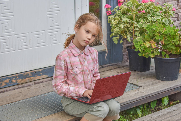 a girl child with a laptop is sitting on the porch of the house during a lesson on a remote photo without a filter - Photo, Image