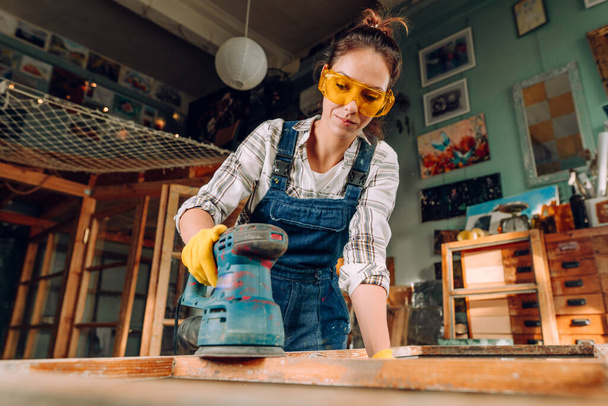 Een portret van een vrouw met een beschermende bril en handschoenen werkend in de werkplaats met behulp van een schijfschuurmachine. Upcycling-concept. - Foto, afbeelding