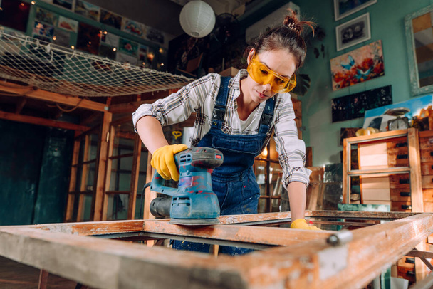 Das Porträt einer Frau mit Schutzbrille und Handschuhen, die in der Werkstatt mit einem Scheibenschleifer arbeitet. Upcycling-Konzept. - Foto, Bild