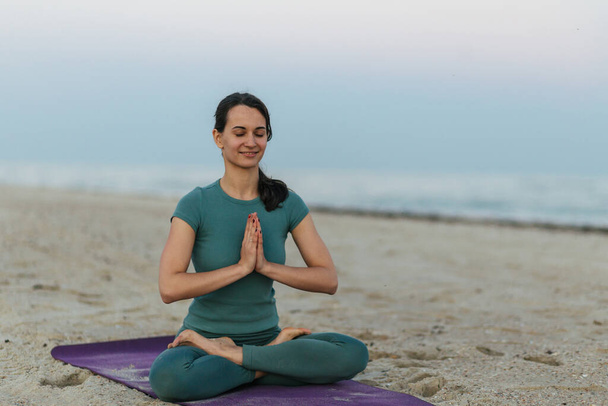 молодая женщина сидит на пляже в позе лотоса и медитирует - Фото, изображение