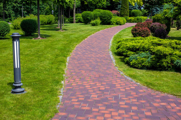 lantern iron ground garden lighting of a park path paved with stone tiles in the backyard among plants, bushes and trees surrounded by a green lawn on a sunny summer day, nobody. - Фото, изображение