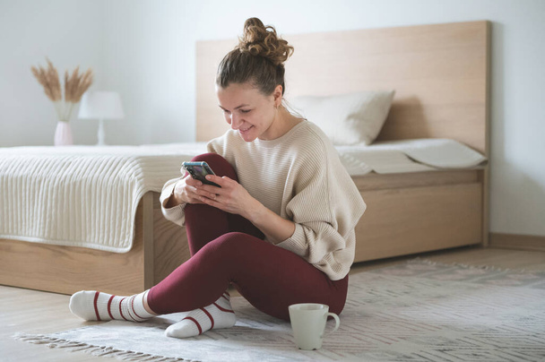 Happy pretty young woman sitting with cellphone on floor in bedroom at cozy home - 写真・画像